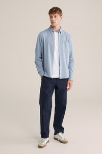 Mlad moški v svetlo modri srajci s kratkimi rokavi nad belo majico, temno modrih hlačah in belih supergah, stoji z rokami v žepih.