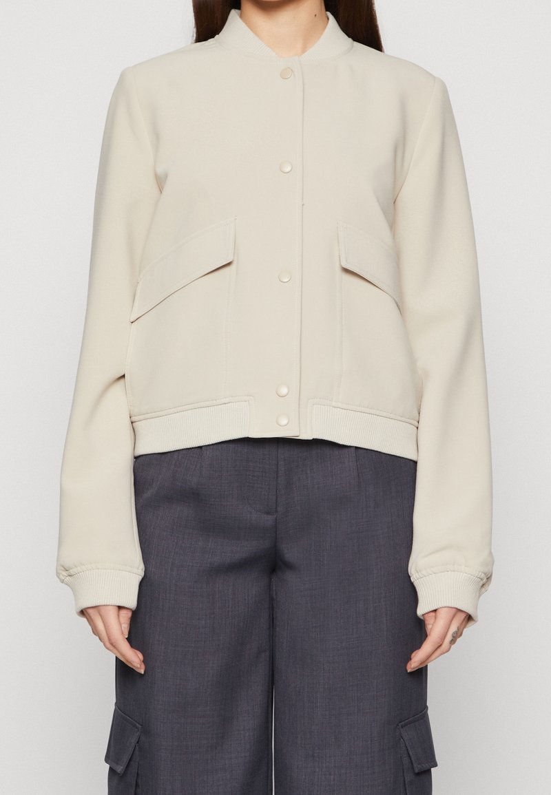 Femme portant une veste crème boutonnée avec de grandes poches avant et un pantalon large gris foncé avec des poches latérales.