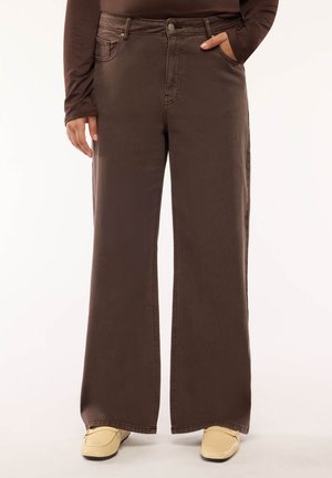 Persona che indossa pantaloni marroni a vita alta e gamba larga, una maglia marrone a maniche lunghe, mocassini beige, e una mano in tasca, in piedi davanti a uno sfondo semplice.