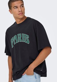 Zwart katoenen T-shirt met een groene "PARIS" afbeelding, losse pasvorm, ronde hals en korte mouwen, gecombineerd met lichtblauwe spijkerbroek.