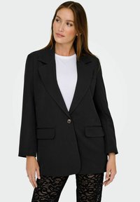 Černý blazer s jedním knoflíkem, klopovým límcem a dvěma předními kapsami. Nosený přes bílé tričko, doplněný černými krajkovými legínami.