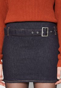 Gonna di denim con una tonalità blu scuro, caratterizzata da un design aderente e una cintura abbinata con fibbia e occhielli argentati. Dettagli di cucitura arancione.