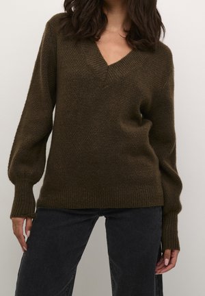 Femme portant un pull texturé col en V vert olive foncé et un jean foncé, debout devant un fond clair uni.