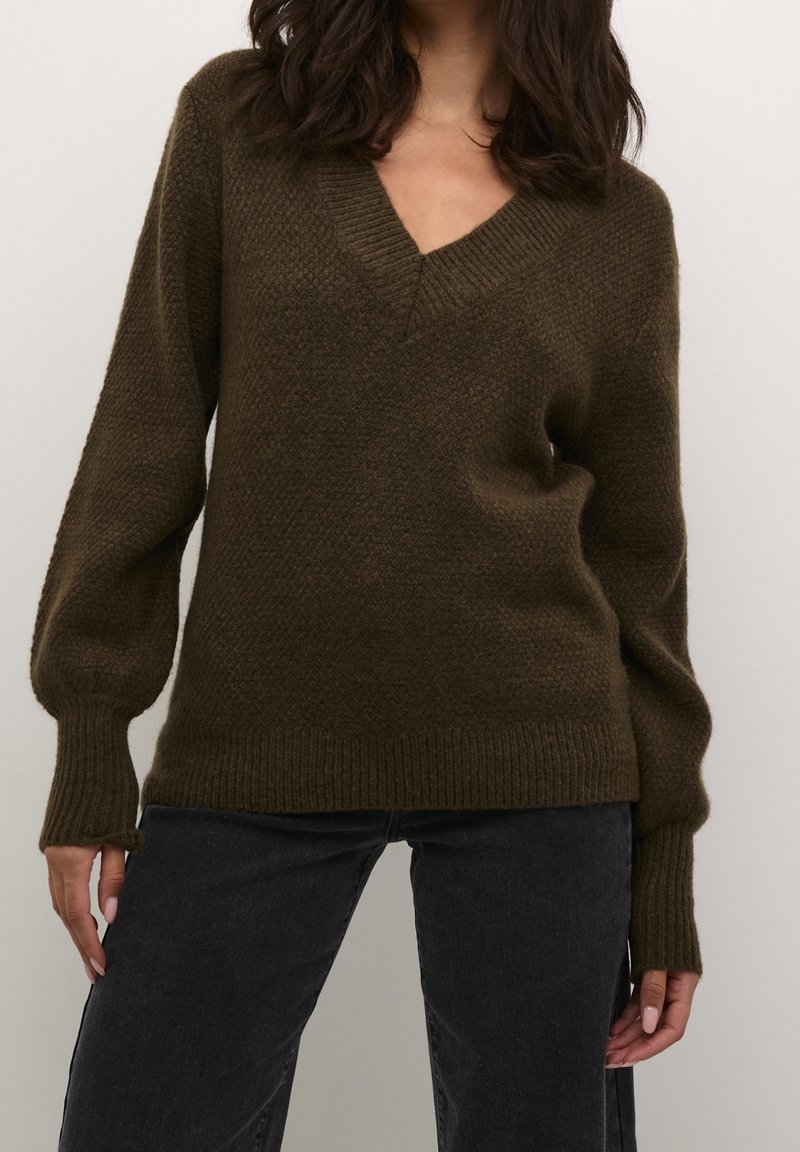 Femme portant un pull texturé col en V vert olive foncé et un jean foncé, debout devant un fond clair uni.