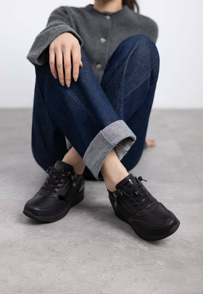 Zapatillas deportivas negras con un acabado texturizado, diseño de cordones y cremalleras laterales, combinadas con jeans de mezclilla arremangados y un suéter gris.