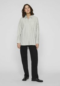 Camisa de manga larga y corte suelto con rayas verticales en verde claro y blanco, con botones en la parte delantera y un bolsillo en el lado izquierdo, combinada con pantalones negros.