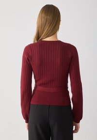 Sweater de manga longa em tom bordeaux, com cintura justa e decote redondo, apresentando um tecido texturizado e um acabamento suave. Vista traseira.