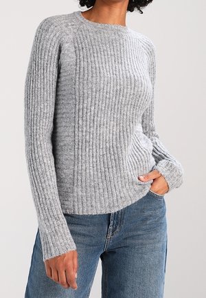 Personne portant un pull côtelé gris clair à manches longues, une main dans la poche d’un jean bleu, sur un fond uni.