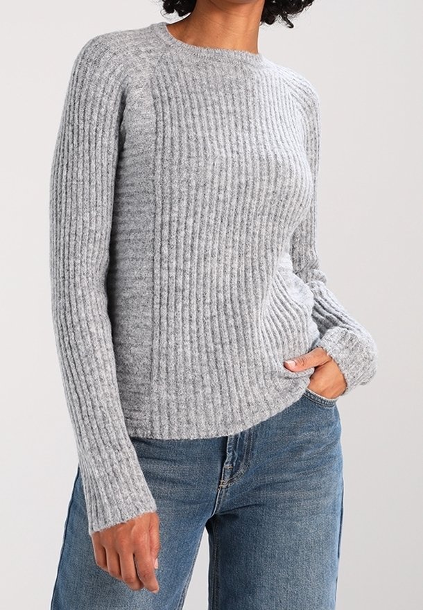 Personne portant un pull côtelé gris clair à manches longues, une main dans la poche d’un jean bleu, sur un fond uni.