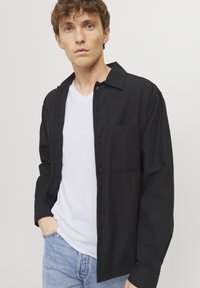 Schwarzes Hemd mit Knöpfen und einer Brusttasche, getragen über einem weißen V-Ausschnitt-Shirt. Kombiniert mit hellblauen Jeans. Glatte Stoffqualität, lockere Passform.