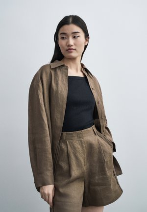 Woman with long dark hair wearing a loose brown shirt, matching high-waisted shorts, and a fitted black top, standing against a plain background.