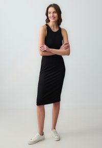 Zwarte mouwloze midijurk met een getailleerde pasvorm, gladde textuur en een ronde halslijn. Gecombineerd met witte sneakers voor een casual look.