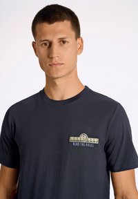 Tummansininen puuvillapaita, jossa on pyöreä kaula-aukko. Etupuolen vasemmassa rinnassa on brodeerattu graafinen kuvasto mittanauhasta ja teksti "Rikko säännöt".