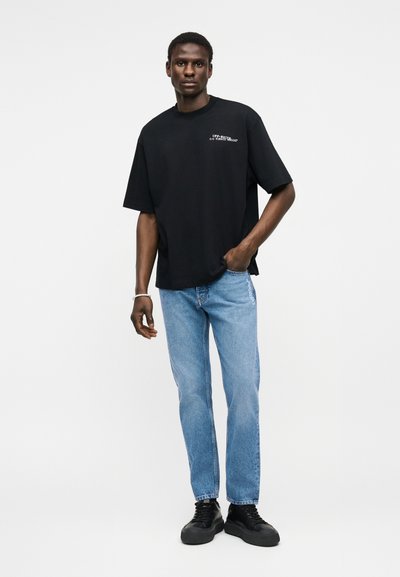 Homme portant un T-shirt noir ample avec un texte blanc, un jean bleu clair avec un texte sur le côté, des baskets noires et un bracelet, debout avec une main dans la poche.