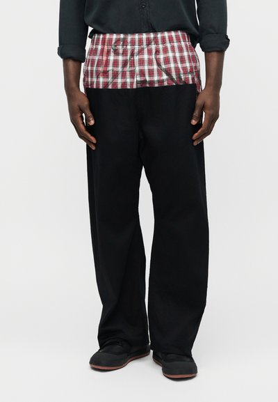 Άνδρας που φοράει μαύρο παντελόνι με φαρδιά μπατζάκια, με κόκκινη και λευκή καρό ελαστική μέση και μαύρα παπούτσια, σε απλό φόντο.