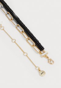 Pulseira dourada e preta com segmentos redondos de borracha, elos de cadeia metálica e um fecho tipo lagosta com uma etiqueta da marca.