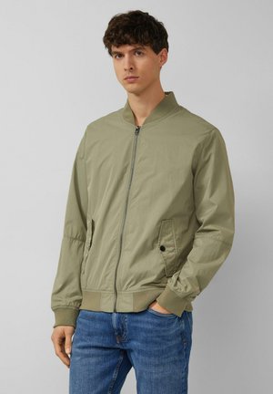 Junger Mann trägt eine hellolivgrüne Bomberjacke mit Reißverschluss vorne, seitlichen Taschen und gerippten Bündchen, kombiniert mit blauen Jeans, steht vor einem grauen Hintergrund.