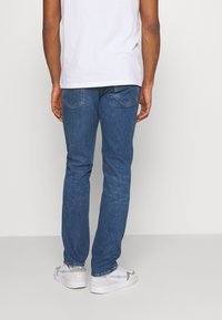 Jeans de mezclilla azul con un corte recto, que cuentan con bolsillos traseros con un sutil diseño de costura. Combinados con una camiseta blanca lisa y zapatos blancos.