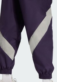 Fialové tepláky majú kontrastný svetlošedý diagonálny pruh, elastické manžety a hladkú, ľahkú textúru látky.