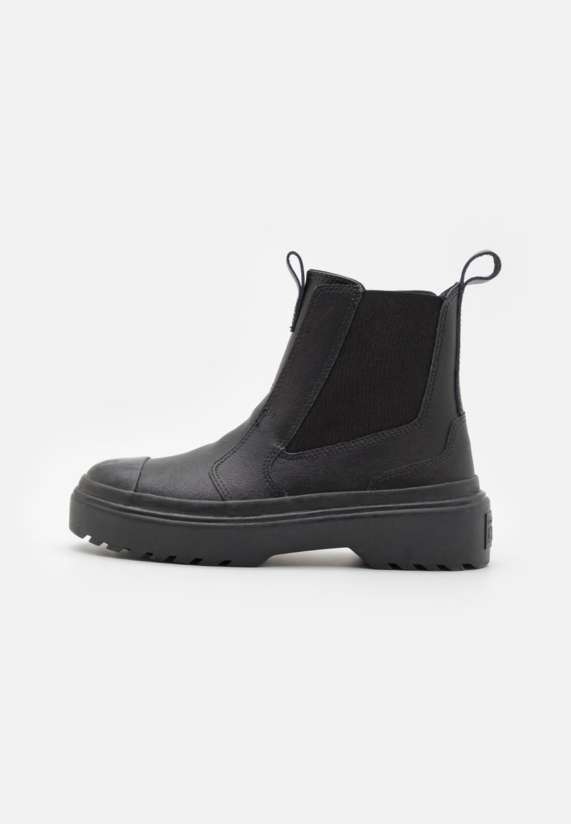 Botte Chelsea en cuir noir avec semelle épaisse, panneaux latéraux élastiques, languettes à l'avant et à l'arrière, montrée de profil sur un fond clair.