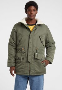 Grüne isolierte Jacke mit einem strukturierten, flauschigen Kragen, vorderen Knöpfen und Zugkordel in der Taille. Verfügt über zwei große Fronttaschen.
