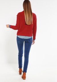 Maglione rosso a coste con scollatura rotonda e camicia di denim blu sotto, abbinato a jeans blu aderenti e stivali alla caviglia marroni.