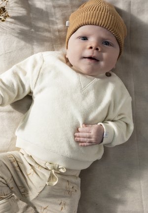 Crème gebreide trui met een v-hals en knoopdetails, gecombineerd met beige broeken met een subtiel gouden patroon. Bruine geribbelde gebreide muts.