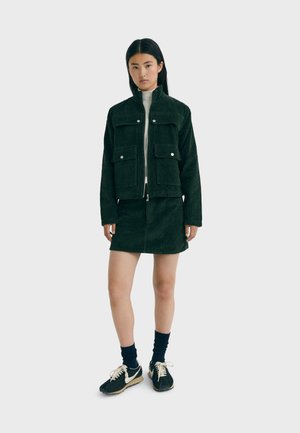Green corduroy jacket with two front pockets, zip-up front, matched with a green corduroy skirt. Model wears black ankle socks and shoes.