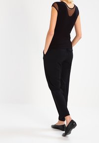 Camiseta negra con detalle de malla en los hombros, combinada con pantalones negros ajustados y zapatos planos negros, vista desde atrás. Textura suave en toda la prenda.