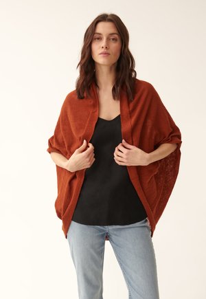 Un cardigan ouvert de couleur rouille en tissu léger, présentant une coupe décontractée avec des manches larges, porté sur un top noir et un jean bleu clair.