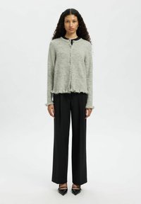 Kobieta o długich kręconych włosach, ubrana w szary sweter na guziki, czarne spodnie z szerokimi nogawkami oraz czarne szpilki z zakrzywionymi noskami, stojąca na białym tle.