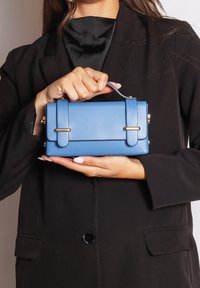 Donna in blazer nero che tiene in mano una piccola borsa rettangolare blu con manico superiore e dettagli in tonalità oro.