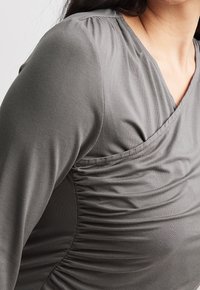 Grijze jersey top met lange mouwen, featuring geplooide details aan de zijkant en een v-hals voor een getailleerd ontwerp. Zachte, gladde textuur.