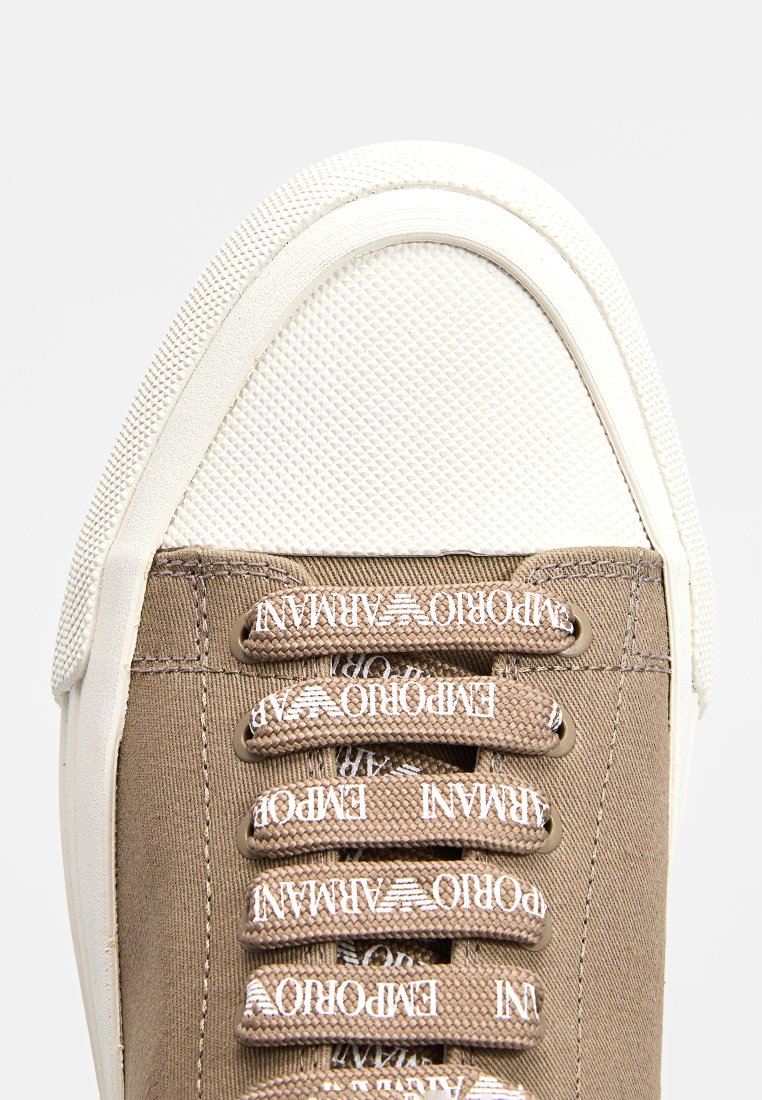 Brauner Leinenschuh mit einer weißen Gummikappe, ausgestattet mit weißen Schnürsenkeln und Ösen. Texturierte Zunge und gebrandete Schnürsenkel mit sich wiederholendem Logo.