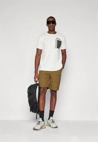 Vīrietis valkā baltu T-kreklu ar krūšu kabatu, brūnas darba stila šortus, melnas zeķes, baltus sporta apavus, tumšas saulesbrilles, rokās tur melnu mugursomu.