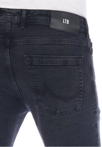 Jeans de mezclilla oscura con una textura suave, que presentan una etiqueta de cuero con marca en la cintura y un bolsillo trasero con un sutil patrón de costura curva.