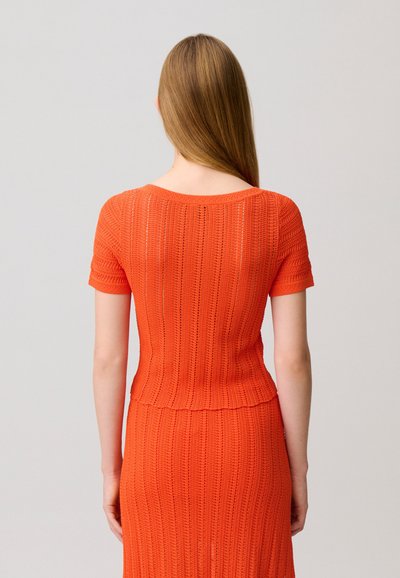 Mujer con cabello liso y castaño, vestida con un top de punto naranja de manga corta y falda a juego, vista desde atrás sobre un fondo liso.