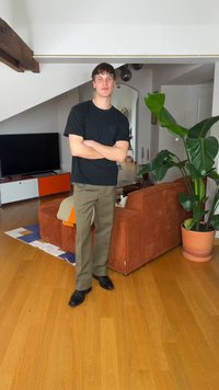 Νέος άντρας με μαύρο πουκάμισο και χακί παντελόνι στέκεται με σταυρωμένα τα χέρια σε ένα μοντέρνο σαλόνι με πορτοκαλί καναπέ και μεγάλο φυτωμένο φυτό.