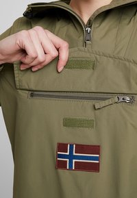 Olivengrøn pullover med lynlås- og velcro-lommer. Har et norsk flag patch, glat tekstur og hætte-design.