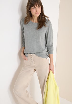 Vrouw leunt tegen een muur, draagt een grijs shirt met lange mouwen, beige broek en houdt een gele jas in haar rechterhand.