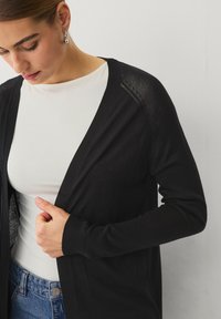 Černý kardigan z lehké, prodyšné tkaniny. Má žebrované manžety a otevřený design, přičemž je nošený přes bílou košili.