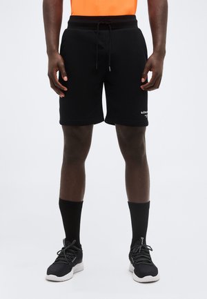 Férfi modell fekete zsinóros rövidnadrágban, fekete középmagas zokniban, fekete-fehér tornacipőben és derékig felvágott narancssárga ingben.