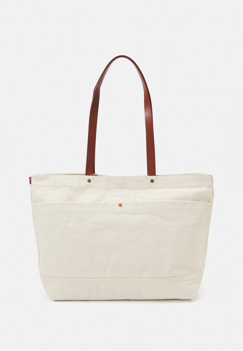 Canvas toteväska i krämfärg, med handtag i brunt läder, rektangulär form, yttre framfickor och diskreta logostickningsdetaljer.