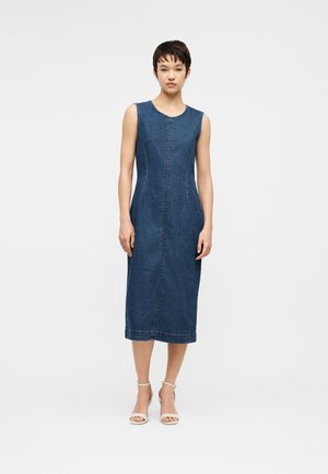 Naine seisab, kandes varrukateta põlvpikkust sinist teksakleiti ja valgeid avatud varbaga kontsakingi ühtlase heleda tausta ees.