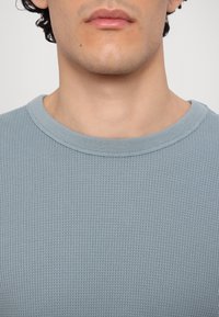 Šviesiai mėlynas, tekstūruotas ilgomis rankedėmis viršus su apvalia iškirpte, turintis vaflinį mezgimo raštą ir raukšlėtą apykaklės detalę.