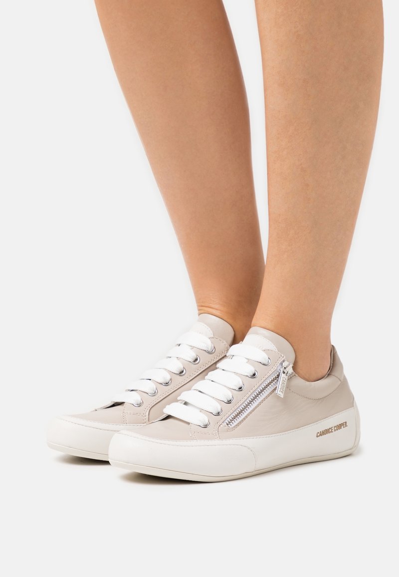 Beige leather sneakers with white accents, featuring a front zipper, rounded toe, and flat rubber sole. Laces in white for additional detail.