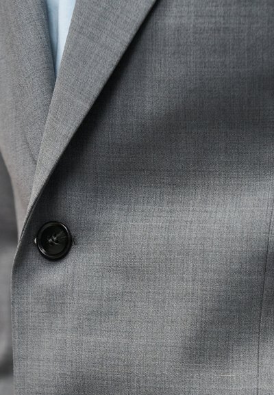 Γκρι σακάκι με μαύρο κουμπί δεμένο πάνω από ανοιχτό μπλε πουκάμισο, που δείχνει το πέτο και το πάνω μέρος του στήθους.