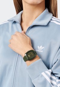 Kvinna som bär en ljusblå Adidas-zipjacka och en olivgrön kvadratisk handledsklocka på vänster handled.