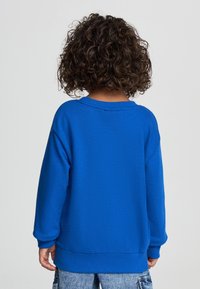 Bambino con capelli ricci che sta in piedi di spalle, indossa una felpa blu brillante e jeans di denim blu chiaro su uno sfondo semplice.