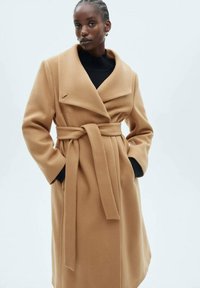 Camel-farbener Wickelmantel aus weichem Stoff mit weitem Kragen, taillierter Taille und schwarzen Innenfutter-Akzenten. Lange Ärmel, mittellanges Design.